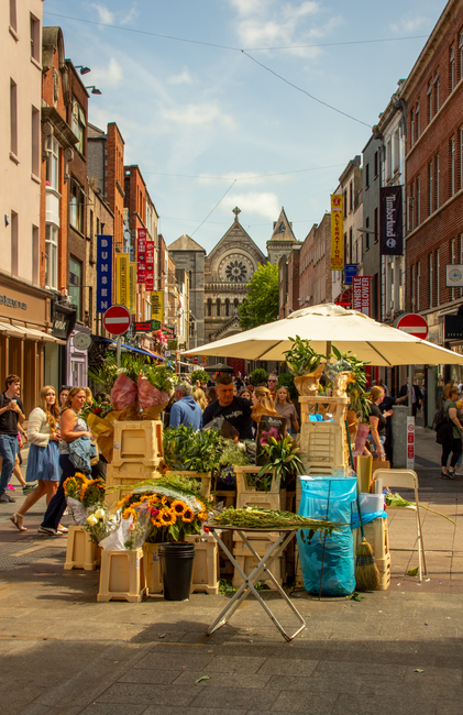 A vibrant outdoor flower market on Stanmore High Street, with a variety of fresh flowers arranged in wooden crates and vases, featuring sunflowers, roses, and greenery. The floral display is set on a
