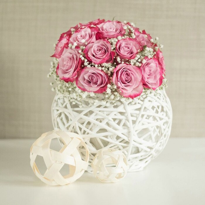 An elegant floral display features a lush, rounded bouquet of pink roses and white baby's breath, styled in a way that would feel at home in a refined living room overlooking the green spaces of Stanmore. The roses, numbering around fifteen or more, are gathered closely into a dome, each one opening in layered petals that shift from a soft, powdery pink in the centre to a more intense, almost fuchsia hue at the outer edges. These two-toned blooms are laced with generous sprigs of gypsophila, whose tiny white flowers create a light, cloud-like veil around the roses and soften the overall silhouette. The arrangement is held in a spherical white vase fashioned from interwoven strands of what looks like painted wicker or rattan, its open, basket-like construction giving the piece a contemporary, airy character. Resting on a spotless white surface that reflects the gentle light, the vase is accompanied by two decorative woven balls in pale natural tones, their strip-like construction forming open geometric shapes that mirror the vase's texture. The background is a warm beige or off-white wall with subtle texture, perhaps reminiscent of soft plaster or fabric wallpaper often found in well-kept homes around Stanmore Hill, providing a quiet, neutral canvas. Soft, diffused lighting enhances the pinks and whites, highlighting the craftsmanship of the bouquet and the interplay of light and shadow through the woven materials. The scene suggests a bouquet ideal for thoughtful gifts, whether for Mother's Day, a birthday, or a romantic surprise, bringing a sense of calm sophistication and gentle celebration into any Stanmore interior.