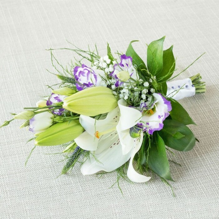 A sophisticated, elongated bouquet stretches diagonally across a light beige, slightly nubby fabric that could be a piece of soft linen laid on a table in a Stanmore home. The bouquet is anchored by several striking white Oriental lilies, their large, trumpet-shaped blooms fully open so that every curve of petal is visible. The petals are creamy white with a smooth, almost satin sheen, gently flaring back to expose warm yellow-orange stamens dusted with pollen and a sprinkling of fine reddish-brown speckles that give each flower its own pattern. Between these open lilies, fresh green buds in a vivid, almost citrus chartreuse stand tall and sleek, hinting at future blossoms and adding a modern burst of colour. Threaded throughout are Lisianthus flowers, also known as Eustoma, their petals gently ruffled and cup-like. They appear in white with dramatic purple edges, some with soft lavender transitions and deeper violet centres, adding both romance and a contemporary twist. Delicate threads of white baby's breath (Gypsophila) scatter across the bouquet, forming a gauzy layer of tiny blooms that soften the more dramatic shapes. The greenery is abundant and thoughtfully chosen: broad, glossy dark green leaves provide structure; finer, airy fronds and a few spikier elements give lift and movement, making the bouquet feel alive rather than static. At the base, the stems are tightly bound with a smooth white satin ribbon, accented with small, pearl-like beads along the thinner end on the right side, evoking the detailing on a bridal sash. Gentle, diffused lighting caresses the flowers, bringing out the texture in every petal and leaf without casting strong shadows. This composition feels ideal for a Stanmore wedding, perhaps photographed before being carried out towards Canons Park, or as an elegant gift arrangement created by a florist who knows how to balance tradition and modern style for local celebrations.