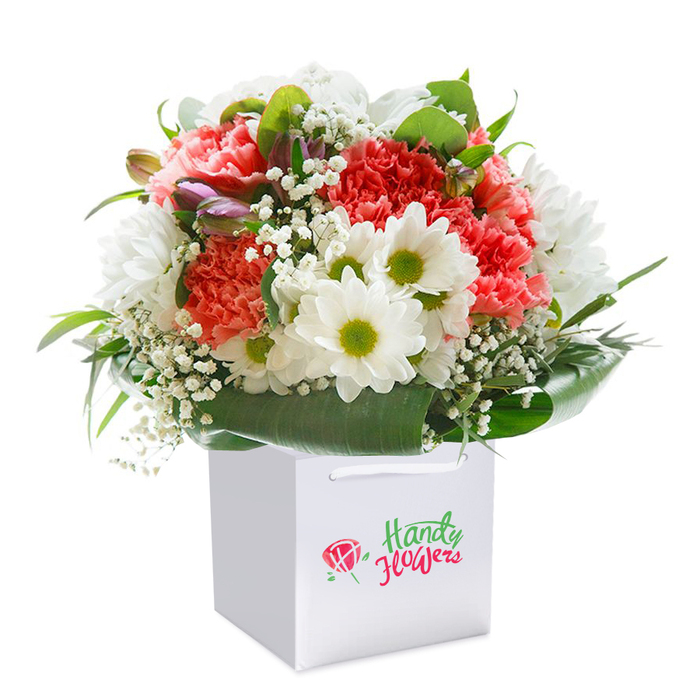 Brimming with artistry and vitality, this floral arrangement sits upright in an immaculate white square gift bag, its colours dancing beneath soft natural light. The main attraction is a vivid collection of coral and pink carnations, their layers of ruffled petals forming a cheerful, textured canvas reminiscent of the cheerful community spirit at Stanmore's weekly markets. Alternating between these are pristine white daisy-like blooms, each displaying a refreshing lime-green centre that pops with energy against the warmer shades. Tiny, clustered stars of baby's breath float throughout, adding breeziness and light contrast-evoking a sense of new beginnings and gentle mornings by Stanmore Common. A subtle touch of purple appears from unopened buds, tucked understatedly within the bouquet, adding intrigue and anticipation. The thriving green foliage, polished and robust, wraps around the bottom edge, its leaves arching like the graceful trees lining Stanmore's elegant avenues. This bouquet's perfectly rounded silhouette ensures every element is given room to shine, while the sturdy white handles of the accompanying bag make it ideal for delivering fresh joy anywhere from Church Road to Stanmore's peaceful residential corners. Imbued with the thoughtful skill and dedication of local Stanmore florists, this arrangement beautifully represents a considered gift for life's many occasions-be it a birthday, anniversary, or simply to show appreciation for someone special in this charming North London suburb.