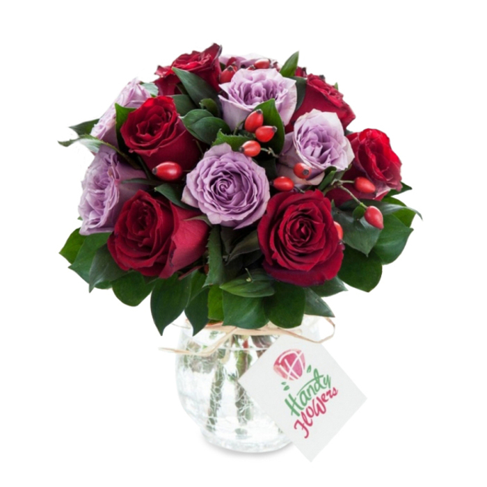 A carefully composed arrangement of velvet-red and soft-lilac roses sits in an antique-effect glass vase, its rounded base tapering into a neat neck, the mercury-like mottling throwing back soft glints of studio light. The bouquet is densely packed, with deep crimson and burgundy roses positioned alongside pale mauve and dusty purple varieties, each bloom freshly opened so the layered petals overlap in a seamless, tactile carpet. Dark green, glossy foliage-probably Ruscus or Salal-frames the blooms and slips between them, creating pockets of cool contrast and an earthy note that complements the roses' sweetness. Interspersed among the petals are small, bright red-orange rose hips whose smooth, jewel-like surfaces add a rustic counterpoint to the roses' plush textures. A natural raffia tie encircles the vase's neck and a rectangular white tag, bearing a minimalist rose motif and green-script naming, hangs forward as a quiet signature of the florist's craft. The whole composition sits against a pure white backdrop that heightens the richness of colour and the subtle reflection in the glass, conveying a mood of romantic luxury and considered gifting. Locally crafted by Flowers Stanmore, this bouquet feels at home both on an elegant dining table and as a delivered surprise for birthdays, anniversaries, or moments when you want to send warmth and style to addresses across Stanmore, Stanmore Hill and the surrounding Harrow neighbourhoods.