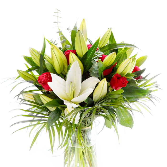 A pristine, studio-style image of a refined bouquet anchored by one large, fully open creamy-white Oriental lily, its smooth petals slightly curved and revealing pale yellow stamens and a soft green pistil at the centre. Around this central flower, a scatter of lime-green and pale yellow lily buds-some tightly closed, others just beginning to give-adds a sense of unfolding life and layered texture. Nestled throughout are rich red rosebuds and early roses, their velvety petals still mostly curled and offering dramatic flashes of passionate colour against the lilies' calm. The bouquet is woven through with varied foliage: deep, forest-green leaves that build structure and shadow; long, feathered palm-like fronds in a brighter emerald that cascade and suggest gentle movement; and silvery-green eucalyptus sprigs with their rounded leaves, which lend a delicate, softened tone. The glass vase is plain and cylindrical, the clear water and visible green stems contributing to an overall feeling of freshness and simple refinement. Against the clean white background, each hue and texture reads clearly-the creamy lily, the lime-tinted buds, the crimson accents, and the layered greens-making this a versatile arrangement suitable for celebrations, anniversaries or as a thoughtful sympathy gift. Imagined for homes around Stanmore Common or for a small dinner in a local Stanmore home, this bouquet radiates quiet elegance; it's the kind of composition a florist would prepare to convey purity and sincere emotion without excess, combining scent, form and colour in careful harmony.