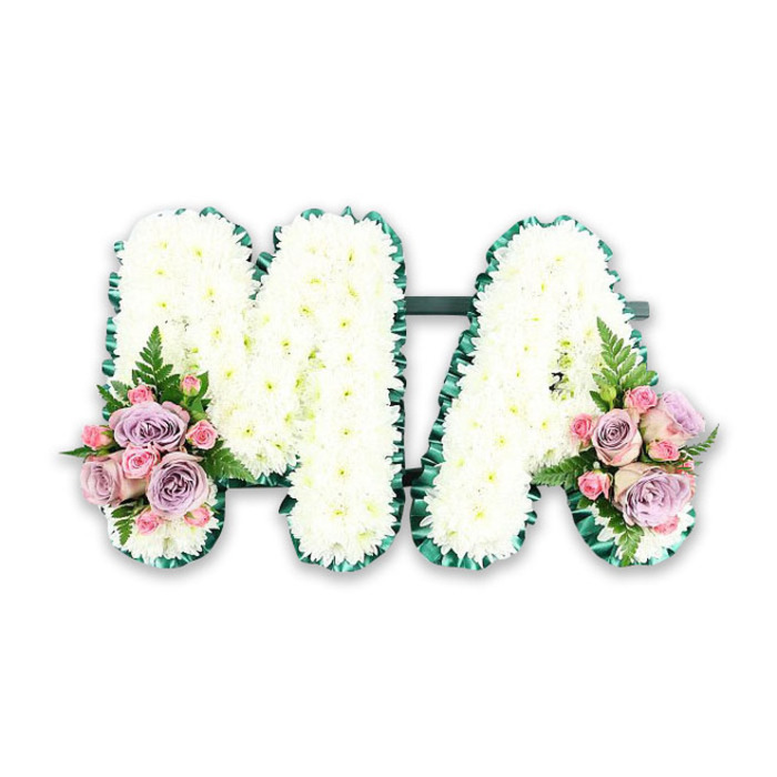 A personalised funeral letter tribute spelling MA is displayed in a simple, well-lit studio setting, letting its thoughtful details speak for themselves. It is the sort of piece a family in Stanmore might choose to commemorate a devoted mother, conveying love and respect through classic, understated floristry. Both letters are formed from sturdy floral bases and are completely blanketed in white chrysanthemums. Each small bloom is positioned closely, their intricate petals creating a soft, textured surface that looks almost like white velvet from a distance. The strong shape of the capital M with its twin peaks and the open, triangular form of the A are kept perfectly legible, their lines crisp despite the softness of the flowers. Running around the edge of each letter is a border of rich green ribbon, gently pleated to contour every angle. This darker frame acts like a line of ink around a drawing, making the design stand out and hinting at the green tranquility of nearby open spaces such as Stanmore Country Park. At the lower left foot of the M and lower right foot of the A, the florist has added delicate sprays of accent blooms. These mini bouquets feature soft pink roses, muted lilac roses, and clusters of pink spray roses, arranged so their rounded heads nestle closely together. Their pastel hues bring warmth and a sense of gentleness to the pristine white base. Slender fronds of fern and touches of deep green foliage extend from behind the roses, adding light, airy movement. This carefully balanced tribute would sit gracefully by the coffin or at the front of a service in Stanmore, offering a quiet yet eloquent message of remembrance for Ma.