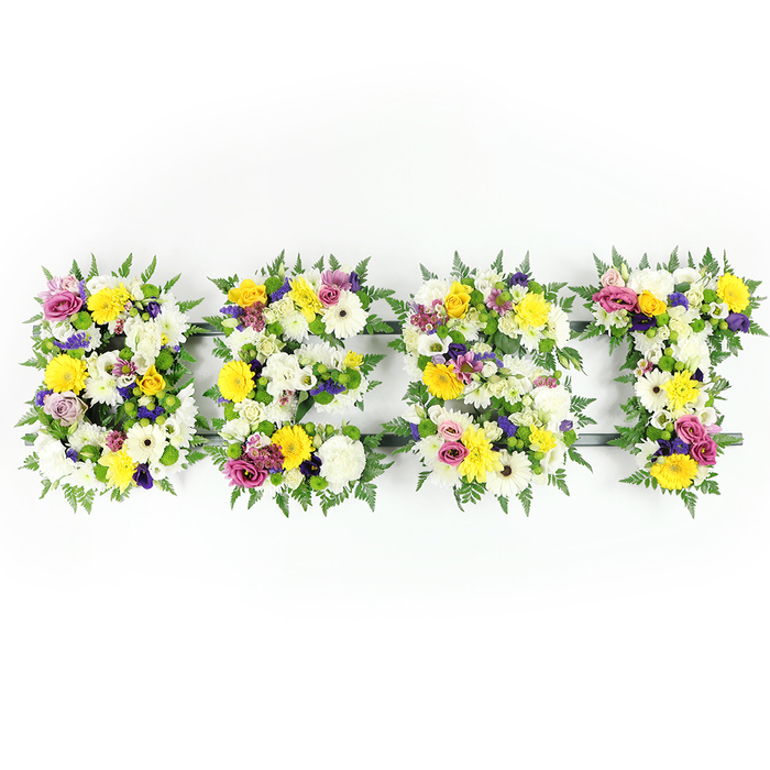 Against a pristine white backdrop, four bold letters form the word BEST, each letter fashioned from a compact, exuberant assembly of fresh flowers and deep green foliage. The dominant tones are pure white and sunny yellow: large, multi-petaled white chrysanthemum-like blooms and ivory spray carnations create a serene, luminous canvas, while bright yellow roses and gerbera daisies provide warmth and a buoyant, celebratory energy. Embedded within the primary palette are clusters of soft lavender and rich purple lisianthus or small chrysanthemums, which introduce a layer of gentle dignity and visual depth, and lively magenta-pink spray roses or carnation-like blooms act as bright, affectionate accents. Dark green fern fronds edge and cradle each letter, their fine textured leaves softening the hard letterforms and lending a graceful, botanical border. Through tiny interstices in the floral mass a slim dark-green or understated metal framework is visible - especially across the E's center bar and the horizontal of the T - a careful reminder of the craftsmanship and support beneath the floral surface. The photograph's even, daylight-balanced lighting enhances the petals' tactile warmth and the glossy sheen of the greenery, making each bloom appear freshly cut and gently hydrated. This arrangement reads as both tribute and celebration, suitable for a funeral service or a commemorative event where families gather to remember a cherished life. I imagine it delivered to a chapel near Stanmore Broadway or laid by loved ones returning up Stanmore Hill, offering comfort, colour, and a floral language of esteem.