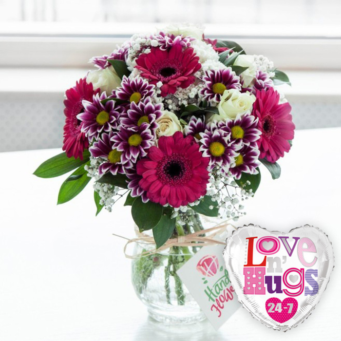 A vibrant, lovingly assembled bouquet stands proudly in a clear glass vase, accompanied by a heart-shaped foil balloon, creating a complete gift moment that would feel at home in any Stanmore living room or office. The bouquet itself is an artful blend of colour and texture, with several large gerbera daisies in a deep magenta-fuchsia hue providing the main visual anchor. Their petals form perfect rings around inky, dark centres, offering a bold contrast that draws the eye. Surrounding these, clusters of smaller chrysanthemums fill the arrangement, their petals transitioning from rich purple at the base to clean white at the tips, each bloom finished with a bright yellow centre that adds a cheerful, sunlit touch. Creamy white roses are scattered gently throughout the bouquet, some still partially closed, lending a soft, romantic quality that balances the vivid daisies and chrysanthemums. Fine sprigs of white baby's breath drift through the design, creating a delicate, airy layer that softens the overall appearance and suggests a light, fragrant presence. Fresh green leaves punctuate the flowers, peeking from behind and between the blooms to add depth and a sense of natural abundance. All the stems are gathered into a rounded, transparent glass vase, where you can see the fresh water and carefully trimmed green stems, a subtle nod to the skill and care behind the arrangement. Around the neck of the vase, a piece of rustic natural twine is tied into a simple bow, holding a small white tag adorned with a pink rose motif and a line of text in a friendly, hand-drawn style, evoking the personal touch of Flowers Stanmore. To the right stands a glossy, silver-edged heart-shaped balloon, its white centre emblazoned with colourful, patterned letters spelling out a loving message and a prominent 24-7, underlining enduring affection. The softly lit, blurred background suggests daylight streaming into a Stanmore home, making this scene perfect for anniversaries, birthdays, or any moment when heartfelt emotion needs to be expressed in flowers.