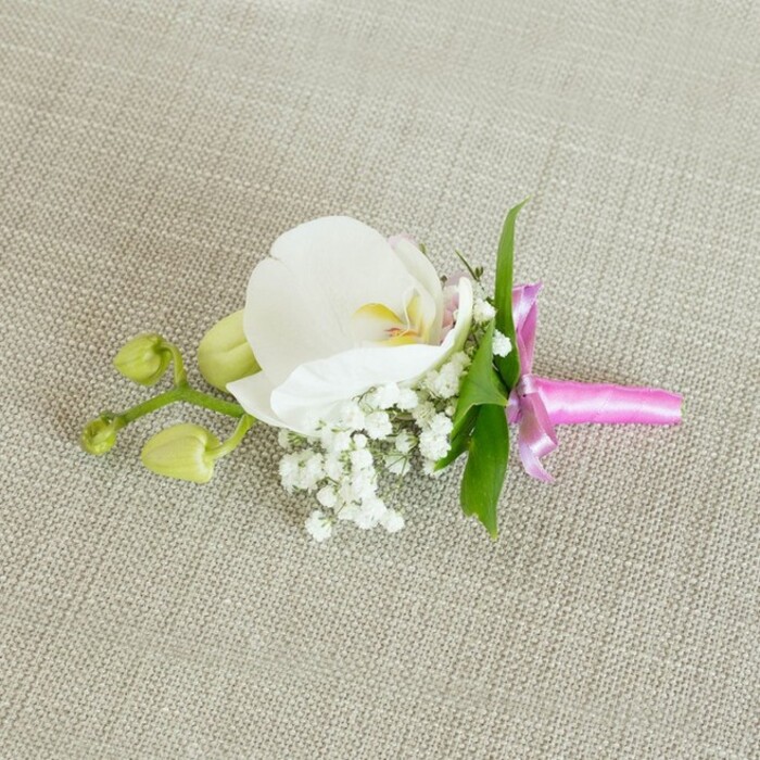 An elegant floral boutonniere is displayed with care on a softly textured, light oatmeal linen surface, reminiscent of the understated, classic interiors of many Stanmore homes where families gather before a big celebration. Placed horizontally across the frame, the arrangement centres on a single, pristine white Phalaenopsis orchid, its broad petals unfurling in smooth, gentle curves. The orchid's throat reveals intricate detail: a soft wash of yellow with the slightest flush of pale pink, adding a hint of warmth and romance to the otherwise cool white bloom. Cradling the base of the orchid is a cushion of delicate baby's breath, each tiny white Gypsophila flower forming a cloud-like cluster that adds softness and a sense of airiness to the composition. Peeking from behind the central bloom are a few slender, vibrant green leaves, their fresh colour echoing the greenery found around Stanmore Country Park and giving the design a natural, garden-inspired touch. Extending to one side is a short stem carrying a couple of tightly closed, lime-green orchid buds, their smooth, rounded forms suggesting new beginnings and the quiet anticipation of blossoms yet to open-perfect symbolism for a wedding, engagement, or prom. The stems of the entire piece are neatly bound together with a bright fuchsia pink satin ribbon, tied into a petite, elegant bow with short, flowing tails. The ribbon's luminous sheen introduces a lively burst of colour that feels celebratory without overpowering the delicate flowers. Soft, even lighting highlights the smooth orchid petals, the tiny rounded blooms of baby's breath, and the subtle weave of the linen background, creating a serene, intimate atmosphere. This timeless boutonniere would sit beautifully on a groom's lapel, a groomsman's jacket, or a special guest's attire at a refined event in Stanmore and the surrounding North West London neighbourhoods.