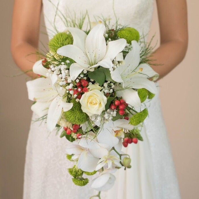 A sweeping, chest-level portrait focuses on a Stanmore bride clutching a breathtaking, cascading bouquet-an arrangement that pays homage to the refined yet lively spirit of the local community. The bouquet boasts several grand white lilies, trumpet-shaped and gently freckled, their stately appearance echoed by the intricate, softly glowing lace of the bride's gown in the background. A classic creamy rose sits confidently at the center, its tightly wound petals suggesting both purity and romance, while clusters of fresh, lime-green cushion mums are woven throughout, adding a zing of contemporary colour and vibrancy, much like spring's arrival in Stanmore's parks. Delicate sprigs of white gypsophila entwine with the main blooms, imparting a dreamy, cloud-like halo. Interspersed hypericum berries, deep red and glossy, provide warm contrast and a celebratory feeling, reminiscent of flower celebrations in the Stanmore village centre. The dramatic silhouette of the bouquet is shaped by a trail of pearly white Phalaenopsis orchids, their flowing form suggesting elegant movement. Long, fine grass blades and deep green leaves curve gently at the bouquet's edges, setting the flowers in a naturally exuberant frame. As daylight caresses each petal, the bouquet resonates with artistry and care-a true testament to the craft of Flowers Stanmore's expert florists and an inspiring centerpiece for a wedding in this leafy north London area.