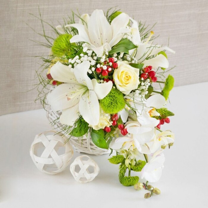 This image presents a sophisticated floral table arrangement that would sit beautifully in a Stanmore home or at a wedding reception near Bentley Priory. At the base, a round white wicker basket anchors the design, its delicate woven pattern creating a light, airy support for the flowers above. Rising from the basket is a full, rounded bouquet of predominantly white blooms. Several large lilies dominate the upper portion, their trumpet-shaped flowers open and slightly tilted at different angles so that their smooth petals and speckled centres are clearly visible. The stamens are softly coloured, suggesting the presence of a subtle, sweet fragrance that might drift across a dining table. Near the top-centre rests a single cream-yellow rose, its petals tightly layered yet beginning to relax, offering a warm, romantic counterpoint to the surrounding whites. From the right-hand side, a string of white phalaenopsis orchids cascades downward, flower after flower arranged in a graceful line. Their broad, velvety petals and delicate throats give the impression of falling water, creating a gentle sense of movement ideal for a bridal top table or an elegant Stanmore dinner setting. The bouquet is richly accented with vibrant green foliage: fine, feathery fronds, bolder glossy leaves, and spherical bright green pom-pom blooms that add unusual, almost moss-like texture. Scattered throughout, tiny white baby's breath blossoms soften the transitions between larger blooms, while small clusters of ruby-red berries offer vivid points of contrast, hinting at winter celebrations or romantic evenings near Stanmore's leafy lanes. The arrangement sits on a clean, white tabletop that reflects faint shadows and highlights. To the left of the basket, two white decorative spheres woven from slender strips echo the basket's texture and provide a modern sculptural balance. The overall effect is one of pure elegance, freshness, and careful craftsmanship, reflecting the refined floral style often created by Flowers Stanmore for local celebrations.
