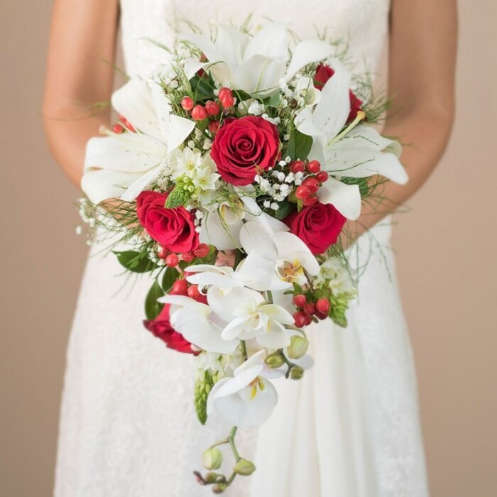Perfect for an unforgettable wedding in Stanmore, this captivating bridal bouquet is held tenderly by a bride in classic white, her elegant gown hinting at lace and romance. The composition is at once modern and timeless-its cascading shape sculpted with striking white lilies, each bloom fresh and alive, evoking the refined beauty of Stanmore's leafy avenues. Deep, luxurious red roses provide a dynamic focal point, their velvety layers intertwined with tumbling trails of white phalaenopsis orchids, whose soft yellow centres and long, elegant stems create a mesmerizing waterfall effect. Sprinkled throughout are clusters of small, vivid red berries that add lively pops of colour, suggesting a festive spirit reminiscent of summer gatherings at Stanmore Common. Gentle clouds of baby's breath and petite white filler flowers infuse the bouquet with softness and movement, echoed in the billowing green ferns and structured foliage forming its base. Bright morning light washes over the bouquet, casting shadows that accentuate every petal and texture while a softly blurred tan background makes the flowers the unmistakable focus. Every flourish-each velvety rose, each fresh green sprig-is expertly crafted, reflecting the tradition, creativity, and care found at Flowers Stanmore, making this bouquet an ideal choice for heartfelt celebrations in and around the area.