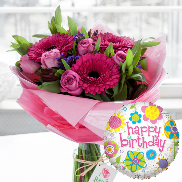 A festive birthday flower and balloon arrangement is shown in a bright, softly focused indoor setting, reminiscent of a sunlit room in a Stanmore family home. Dominating the composition is a clear cylindrical glass vase holding a full, expertly arranged bouquet. The stems, neatly cut and aligned, are partly veiled by layers of translucent light-pink cellophane that flare gracefully around the base of the flowers, creating a gentle, cloud-like frame. At the centre of the bouquet, several large gerbera daisies in vivid shades of fuchsia and magenta command attention, their finely textured petals radiating from deep, velvety centres so dark they appear almost black. Nestled between these bold blooms are numerous soft pink roses, from tiny closed buds to more open flowers whose spiralling petals suggest tenderness and celebration. Threaded throughout are sprigs of small, deep purple filler flowers, adding a cool, shadowy depth that balances the bright pinks, while an abundance of fresh green foliage provides structure and a sense of just-cut garden freshness, like flowers gathered after a stroll near Bentley Priory Nature Reserve. Around the neck of the vase, rustic twine is tied in a simple bow, securing a small off-white gift tag printed with a refined rose motif, subtly signifying a considered, high-quality gift choice. In the foreground to the right, overlapping slightly with the bouquet, a glossy round Mylar balloon adds a playful touch. Its white background, softly flecked with grey, is scattered with whimsical flowers and circular shapes in bright yellow, sky blue, lime green, bubble-gum pink and rich purple. Across the centre, the phrase happy birthday appears in lively pink script, instantly clarifying the occasion. The softly blurred background keeps all attention on the bouquet and balloon, capturing a moment of joyful anticipation, as if this arrangement has just been delivered to mark a special Stanmore birthday.
