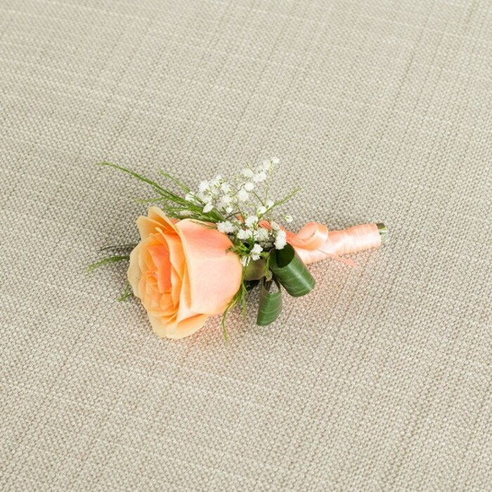 A finely detailed boutonniere featuring a single peach rose is photographed lying horizontally across a light beige, linen-like fabric, the fine weave of the material forming a gentle grid that adds texture without drawing attention away from the flowers. The rose is the undeniable focal point, positioned slightly off-centre and angled so that its spiralling heart is visible, the petals transitioning from a warm, sunlit orange-peach in the core to a softer, creamy pastel at the outer edges. Nestled close to the rose, on its upper side, clusters of small white baby's breath blooms create a lacy frame, their tiny rounded petals giving an airy counterpoint to the rose's smooth, layered curls. Wispy, grass-like green foliage extends from behind the main flowers, adding height and movement, while two prominent, glossy dark green leaves are wrapped partially around the bound stems, providing both structural support and visual depth. The stems are meticulously wrapped in a peach satin ribbon that mirrors the rose's colouring, the ribbon smooth and snug, finished with a petite, tidy bow that sits neatly at the base. Gentle, even lighting washes over the arrangement, enhancing the delicate shadows within each petal and catching the faint sheen of the ribbon while leaving the linen background softly muted. The scene feels calm and composed, reminiscent of a groom preparing in a quiet Stanmore townhouse near Canons Park before stepping out to meet guests. It suggests a special occasion steeped in affection and anticipation, where this small floral detail becomes a cherished part of the day's memory, perfectly suited for weddings, proms, or formal celebrations across Stanmore and the surrounding North West London neighbourhoods.