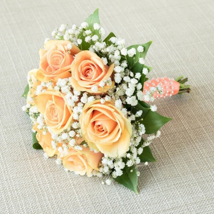 Perfectly placed against a softly textured beige backdrop, this hand-tied bouquet crafted by skilled Stanmore florists lies at a gentle angle, echoing the tranquil landscape of Stanmore Country Park. At its heart, seven lush, open peach and pale apricot roses capture the essence of warmth and celebration, their petals elegantly unfurling from the center in spirals, each bloom radiating poise and vitality. Interspersed among the roses are abundant tufts of white baby's breath, creating a featherlight, cloud-like effect-a visual echo of the morning mist that often settles over Stanmore's leafy streets. Hidden beneath and between the blooms, clusters of deep green foliage peek forth, lending the structure and rich contrast characteristic of the area's beloved private gardens. The stems are tightly gathered and wrapped in a soft peach ribbon, lending the bouquet its distinctive bridal finish, while a spiral ribbon of polished white pearls brings an extra note of refinement, reminiscent of the delicate details adorning Stanmore's historic wedding traditions. Soft, harmonious lighting bathes the bouquet, giving prominence to each petal's texture and every pearl's gentle shimmer. This composition would be at home among wedding festivities in Stanmore, near the halls of Bentley Priory, or celebratory gatherings around the area's beautiful open spaces-a bouquet that celebrates love stories as timeless as the community in which it was created.