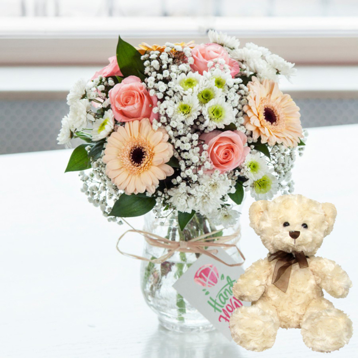 Set against a bright, softly blurred background, this anniversary gift composition rests on a pristine white surface, much like a well-lit kitchen table in Stanmore on a sunny afternoon. In the centre stands a clear glass jar-style vase, its simplicity allowing the pastel bouquet it holds to take full attention. The arrangement is a gentle blend of soft pink roses, their petals layered and romantic, and peach gerbera daisies with dramatic dark brown centres that form a cheerful focal point. Nestled around them are petite white chrysanthemums, some with whispers of lime-green at their centres, lending freshness and volume. Threaded throughout, tiny white baby's breath adds a lace-like delicacy, softening the edges of the design and creating a sense of airiness. The stems, visible through the glass and resting in clear water, are neatly gathered, speaking to the careful craftsmanship behind the bouquet. Around the vase's neck, a piece of natural twine is tied in an unfussy bow, and from it hangs a small rectangular white tag. The tag is adorned with a pink heart and graceful green script, a visual cue that this is a thoughtfully curated, possibly handcrafted gift, perfect for celebrating years shared together.
To the right of the vase, a cream-coloured teddy bear sits upright, positioned as if keeping close watch over the flowers. Its plush fur appears soft and slightly textured, inviting touch. Dark bead eyes and a small brown nose give it a gentle, endearing expression, while a narrow dark brown satin ribbon tied in a neat bow around its neck echoes the deeper tones of the gerbera centres. The overall lighting is bright yet soothing, with the out-of-focus background ensuring no distractions from the intimate pairing of blooms and bear. This peaceful, affectionate scene would be right at home in a Stanmore living room, marking an anniversary with quiet joy and a keepsake that lingers long after the flowers have faded.