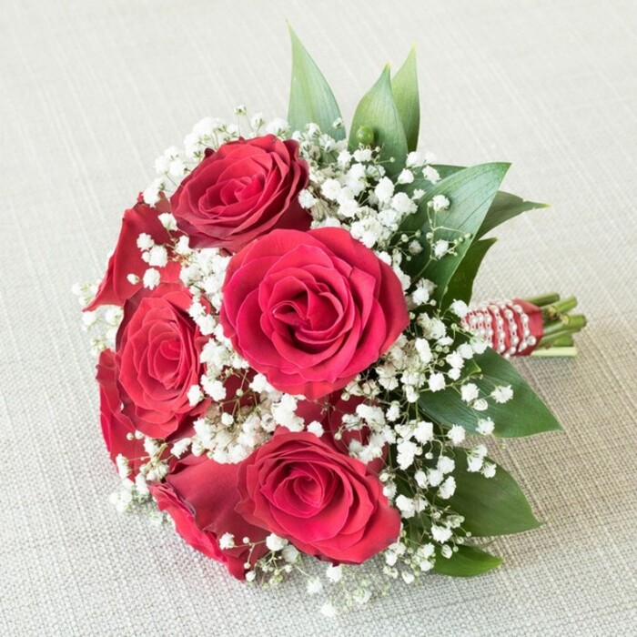 Elegantly displayed against a soft beige textured linen, this classic hand-tied bouquet from Flowers Stanmore radiates sophistication and heartfelt sentiment, making it a favorite in wedding parties across Stanmore and beyond. Central to the display are six meticulously chosen red roses, each one opening from a tightly furled bud to reveal layer upon velvety layer of sumptuous crimson-a dramatic focal point set off exquisitely by bands of delicate white baby's breath (Gypsophila). These frothy accents tumble playfully between the roses, casting a gentle, airy frame that transforms the vivid reds into a romantic daydream. Supporting the composition, lush green leaves-perhaps ruscus or salal gathered from trusted local suppliers-peek out, lending structure and freshness, reminiscent of Stanmore's leafy avenues. The bouquet's stems are precisely bound with a deep red satin ribbon, skyward-wrapping a spiral of small, radiant white pearls, evoking a sense of refined luxury. Bathed in even, natural light, every petal and pearl gleams softly, while the overall arrangement conveys themes of love, new beginnings, and timeless tradition-apt for wedding articles, anniversary gift ideas, or stories celebrating Stanmore's flourishing floral artistry. The image captures not just flowers, but the craftsmanship and care that Flowers Stanmore brings to every community celebration.