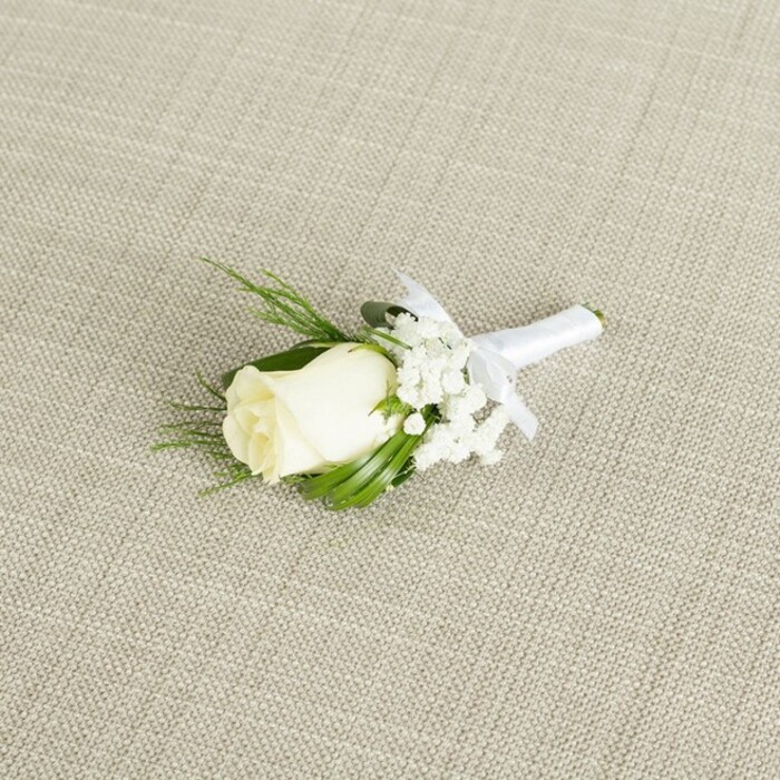 A refined white rose boutonniere rests diagonally across a softly textured, light beige fabric that resembles natural linen, its gentle crosshatch weave visible and inviting to the touch. In the centre of the composition, a single creamy white rosebud is tightly furled, its petals smooth and velvety, forming a classic, conical shape that speaks of freshness and care. The outer petals curl ever so slightly, creating delicate shadows and highlights that the soft, diffused lighting gently emphasises. Nestled to one side and peeking from behind the bloom are gossamer clusters of white baby's breath, their countless petite blossoms forming a cloud-like surround that softens the overall look. Interspersed around the rose and gypsophila are slender, vivid green leaves and a few fine, fern-like stems, adding both structure and organic movement. These varying greens frame the rose, echoing the quiet greenery found around Stanmore's residential streets and the edges of Canons Park. The stems of the rose, filler flowers, and foliage are gathered neatly and wrapped in smooth, pure white satin ribbon. The ribbon is wound tightly and evenly, then finished with a small, neat bow just beneath the floral head, making it immediately clear that this is a boutonniere intended for a lapel-perhaps for a groom exchanging vows in a local Stanmore venue or a smart guest attending a formal event nearby. The neutral background and gentle lighting create a serene atmosphere, allowing the pure whites and greens to stand out without harsh contrast. Crafted with the care of a seasoned florist, this boutonniere exudes timeless sophistication, perfectly suited to weddings, proms, or black-tie occasions across Stanmore and the surrounding North West London neighbourhoods.