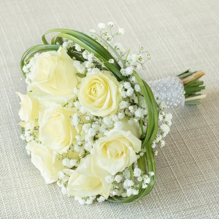 Captured atop a subtly woven pale beige linen, this exquisite bridal bouquet perfectly encapsulates the grace and romance of a Stanmore celebration. Seven or eight generously sized, creamy white roses anchor the arrangement, their petals gently opening from the centre in cascading stages, radiating a sense of poise and delicacy. Artfully interspersed between these roses are delicate sprays of baby's breath, their minuscule white flowers forming soft, floating clouds that echo the peaceful charm of the nearby Stanmore Country Park. Slender, vivid green bear grass is meticulously threaded among the blooms, forming sweeping arcs that frame and elevate the ensemble with a modern flourish. The bouquet's stems are bound together with a shimmering, light grey satin ribbon that wraps securely and is gracefully embellished with several rows of lustrous white pearls, enhancing the bouquet's tactile and visual appeal. The stems' natural ends peek below, giving a glimpse of the fresh greenery and underscoring the bouquet's artisanal craftsmanship. Suited for weddings or anniversaries in Stanmore's storied halls or serene gardens, this floral composition exudes purity, timelessness, and an understated luxury that only a skilled, local florist could create. Its faint, harmonious fragrances linger as a subtle reminder of joy and anticipation, making it a cherished part of any memorable occasion.