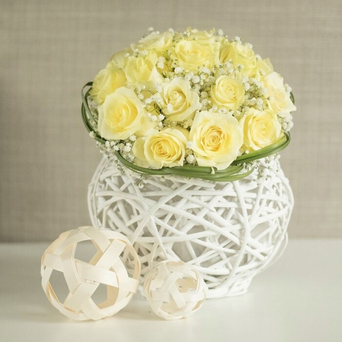 An artfully composed wedding-style centerpiece dominates the scene, radiating quiet luxury that would feel right at home in an elegant Stanmore reception venue. A rounded cluster of pale yellow roses crowns the arrangement, each bloom at its prime, with full, velvety petals unfolding in generous, symmetrical layers. Their colour is soft and creamy, somewhere between ivory and warm butter, creating a cohesive, calming palette. Interspersed among the roses are airy sprigs of white baby's breath, the Gypsophila forming light, cloud-like tufts that nestle between the larger blooms. These tiny white stars add subtle texture and give the piece a romantic, almost ethereal softness. At the base of the floral dome, a slender band of deep green leaves encircles the bouquet like a living ribbon, carefully positioned to give structure and definition. The arrangement is cradled in a distinctive white woven vase made from slender, interlaced strands that create an open lattice pattern. Its shape is gently curving, narrowing slightly through the middle before widening at the top and bottom, like a modern interpretation of an urn. Through the gaps in the weave, soft shadows fall onto the glossy white table surface beneath, which catches a subdued reflection of the flowers and the vase. In the foreground, two decorative spheres of different sizes rest thoughtfully, crafted from pale, natural material woven into open, geometric orbs that echo the vase's texture. The larger ball sits slightly in front and to the left, while the smaller one nestles just beside it, adding a sense of depth and balance. The background is a softly blurred, neutral-toned wall that hints at a calm, tastefully designed interior space. Altogether, the composition suggests joy, grace, and careful craftsmanship, ideal for a beautifully laid table in a Stanmore home or a nearby wedding venue.