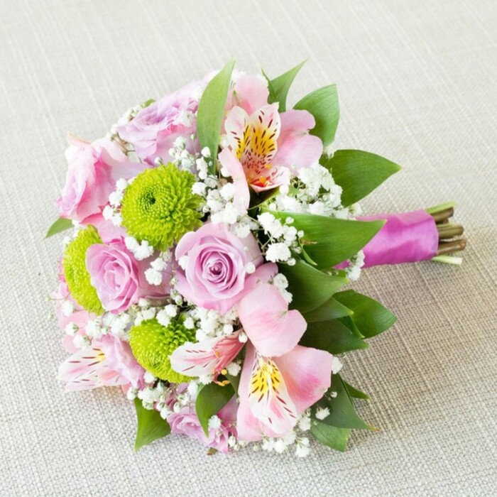 A beautifully composed, hand-tied bouquet lies on a diagonally placed swathe of pale beige fabric, its subtle weave reminiscent of fine linen often seen in cosy Stanmore homes near Canons Drive. The flowers are arranged horizontally, allowing every bloom to be visible in a harmonious blend of pastel pinks, soft lilacs, bright greens, and pure whites. At the centre, pastel lavender-pink roses open in graceful spirals, some with tight, promising buds and others with fully unfurled petals that suggest a gentle, classic fragrance. Around them sit vivid lime-green button chrysanthemums, perfectly rounded and densely petalled, their chartreuse tones injecting freshness and a slightly playful edge to the otherwise romantic palette. Interlaced through the roses and chrysanthemums are pale pink alstroemeria, or Peruvian lilies, their petals slender and gently flared, each bloom revealing a pattern of yellow flashes and fine, dark speckles that add an exotic, detailed charm. Fine sprays of white gypsophila drift through the arrangement, filling small pockets of space and creating a soft, misty halo that ties the colours together and makes the bouquet feel cloudlike and abundant. Lush, dark green foliage frames the composition, some leaves arching outward to elongate the silhouette, others nestled inside to support the blooms and provide a rich contrast to the lighter tones. The stems are gathered tightly at one end and finished with a lustrous magenta-purple satin ribbon, wrapped neatly into a comfortable handle while allowing the trimmed green and brown stem ends to remain visible for a natural, garden-fresh feel. The overall mood is one of springtime romance and joy, perfect as a Stanmore bridal bouquet, a special anniversary gift, or an elegant centrepiece for an intimate celebration at home.