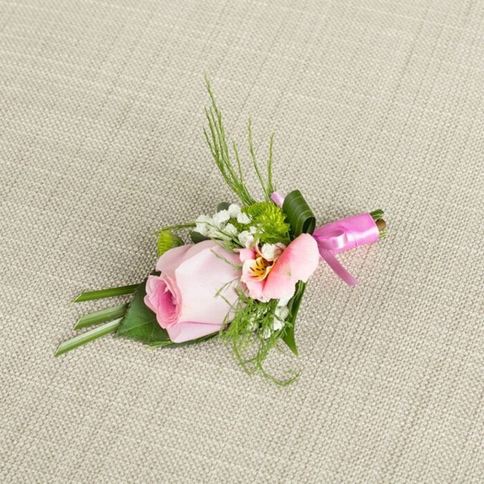 Diagonal on a neutral, textured linen reminiscent of Stanmore's classic style, a bespoke boutonniere shines with sophisticated detail and freshness. The design is centered around a pastel pink rose, tightly curled and full of promise, its inner petals blushing vibrantly before fading to paler edges-a soft echo of spring mornings. Nestled beside the rose, a warmly toned Alstroemeria, with peach-pink petals opening to a heart of yellow and subtle dark accents, catches the eye and enhances the bouquet's visual harmony. Baby's breath forms delicate halos that give the arrangement an ethereal, floating quality, while textured green mums introduce lively pops of colour and a sense of joyful abundance seen in Stanmore's flourishing gardens. Wispy blades of grass and airy fern fronds give height and gentle structure, set against bold, broad leaves folded with care-a reflection of skilled florist artistry. Completing the ensemble, a glossy fuchsia pink satin ribbon binds the stems, tied in a crisp, uplifting bow that glimmers softly under ambient light, ready for ceremonies at Stanmore's historic venues. Every component, from the fresh florals to the tactile greenery, is composed with an eye for balance and local flavour, making this boutonniere an ideal choice for grooms, prom goers, or anyone seeking a refined floral accent for a cherished event in Stanmore.