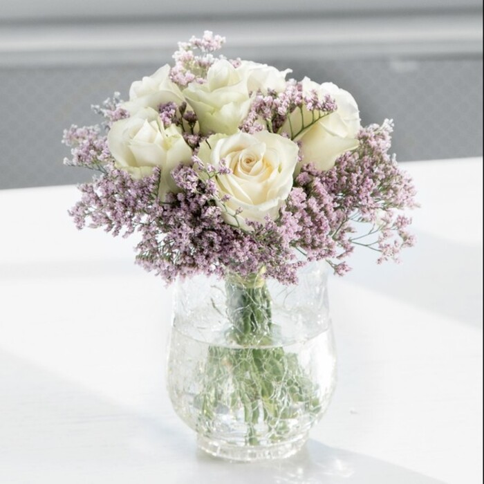 A luxuriously full bouquet of creamy white roses surrounded by airy lavender statice is captured in the centre of the image, its rounded silhouette perfectly balanced and quietly impressive. The roses, likely around a dozen in total, are in that ideal stage between bud and full bloom, their petals opening in soft spirals that create a plush, layered effect. Their colour sits gently between ivory and warm cream, giving the bouquet a timeless, classic beauty often favoured for weddings and anniversaries in and around Stanmore. Nestled among these larger blooms, delicate sprays of lilac-purple statice weave through the arrangement, their many tiny flowers forming a gauzy haze that frames and cushions each rose. The fine, papery texture of the statice adds a whisper-light contrast to the smooth, velvety petals of the roses, lending the bouquet depth without overwhelming its simplicity. All the stems are gathered into a tall, sleek, clear glass vase, the cylindrical shape offering a clean, modern line. Within the vase, straight green stems are visible, some gently bending as they rest in clear water that catches the light, adding a cool, transparent shimmer. The vase stands on a pristine white surface, perhaps a kitchen island in a contemporary Stanmore apartment or a reception counter near Stanmore Broadway, and the white backdrop subtly reflects the vase's outline. The background is softly blurred but bathed in natural daylight, probably from a large nearby window, giving the impression of a bright, tranquil room. This soft light highlights the roses' gentle curves and the statice's tiny blooms, creating an overall atmosphere of calm elegance and quiet affection, ideal for housewarmings, sympathy tributes, or simply brightening a loved one's day. The bouquet feels freshly arranged by an experienced florist, every stem deliberately placed to achieve a sense of poised, modern grace.