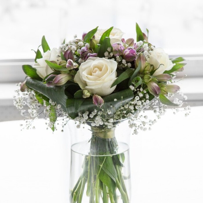 The photograph shows an elegant, rounded bouquet presented in a clear cylindrical glass vase, illuminated by soft, natural light that recalls a bright but peaceful morning in Stanmore, perhaps near the open spaces around Bentley Priory Nature Reserve. At the centre of the design are several pristine white roses, fully open, their layered petals unfurling in generous spirals that feel plush and inviting. They sit like luminous focal points, their gentle ivory tones reflecting the surrounding daylight. Woven through and around these roses are numerous stems of lavender and muted purple alstroemeria, some still in elongated, tightly wrapped buds, others partially open to reveal their slender petals and fine, darker streaks of colour near the throat of the flower. These lilac and soft violet shades lend a subtle but distinct contrast to the creamy whites, creating a calm yet sophisticated palette. Between the larger blooms, airy tufts of white baby's breath drift like tiny stars, softening the transitions and giving the bouquet an almost weightless quality. Encircling and supporting the flowers is an abundance of green foliage, featuring glossy, broad leaves alongside slimmer, arching stems that add depth and gentle movement, as though the arrangement has been freshly gathered from a well-tended garden. The stems are clearly visible through the vase, neatly cut and submerged in clear water, forming an organic pattern that underscores the natural beauty of the flowers. Overall, the bouquet has a carefully sculpted, dome-like silhouette, conveying a sense of order, care, and craftsmanship. The mood it evokes is one of refined comfort and hopeful serenity, suitable for expressing admiration, offering quiet condolence, or celebrating an intimate occasion in a Stanmore home, from modern apartments near the station to traditional houses tucked away off Old Church Lane.