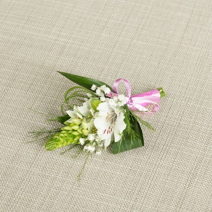 Set against a subtly textured, light beige cloth that resembles finely woven linen, a small yet sophisticated boutonniere draws the eye with its well-balanced mix of white, green, and pink. At its centre, a single white alstroemeria bloom opens fully, the petals wide and slightly ruffled, their surface almost satiny in the soft light. Near the throat of the flower, delicate reddish-brown speckles form irregular streaks, adding depth and a painterly quality that guides the gaze toward the central stamens. Immediately to the left of the main blossom, a slender cone of vivid lime green buds stretches upward, each bud compact and nested tightly against the next so that the cluster resembles a miniature floral spire. This green spike introduces a strong, sculptural line that contrasts beautifully with the softness of the alstroemeria, evoking the fresh, upright growth of plants seen in Stanmore's small front gardens and along its quieter side streets. Threaded throughout the boutonniere, fine stems of white baby's breath appear almost weightless, their tiny round blooms drifting between the larger elements like pockets of soft mist, adding a romantic haze and lightening the overall silhouette. The base of the design is made of lush foliage: broad, dark green leaves form a solid, reassuring backdrop, while slender, flexible blades of bright green grass curve playfully, giving the arrangement a sense of gentle, natural movement. On the right side, the stems are gathered neatly and secured with a glossy satin ribbon in a clear, bright pink hue, tied into a small, tidy bow that introduces a cheerful, celebratory accent. The lighting is even and calm, revealing every subtle vein and petal edge, and the entire piece feels freshly made, perfect for pinning to a lapel at a wedding, evening event, or school prom in the Stanmore area, where understated elegance and personal touches are cherished.