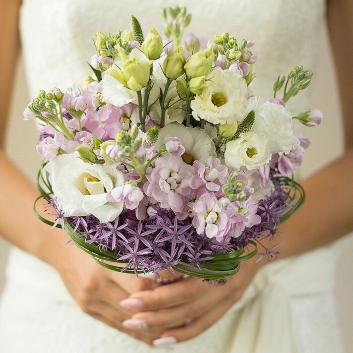 Holding centre stage in this evocative close-up is a bridal bouquet that seems to encapsulate both the refined artistry and natural splendor of Stanmore. The arrangement radiates outward with a lush, rounded harmony-elegant white Lisianthus blossoms, their petals softly ruffled and radiant, are delicately interspersed with unopened greenish buds, symbolizing promise and continuity. Surrounding these focal blooms, light pink and gentle lavender stock flowers tumble in abundance, their multi-petaled forms evoking the tender wildflower meadows found near Stanmore Common at dawn. Along the base, a dramatic collar of deep purple, spiked Allium grounds the bouquet, bringing intriguing texture and visual contrast reminiscent of ornamental borders at Bentley Priory. Artfully looping between the stems and blooms, slender strands of vibrant green bear grass lend a modern, architectural element, echoing the manicured greenery lining Stanmore's residential lanes. The bouquet is carefully supported by a bride's hands, manicured and subtle in nude pink-every detail thoughtfully placed for balance and beauty. In the background, the diffused light and hint of an ivory, lace-embellished dress create an aura of gentle romance, ensuring the bouquet remains the central spotlight. Perfect for an elegant wedding or a feature on floral design trends, this bouquet captures the timeless appeal and refined contemporary style that make Stanmore's celebrations so cherished.
