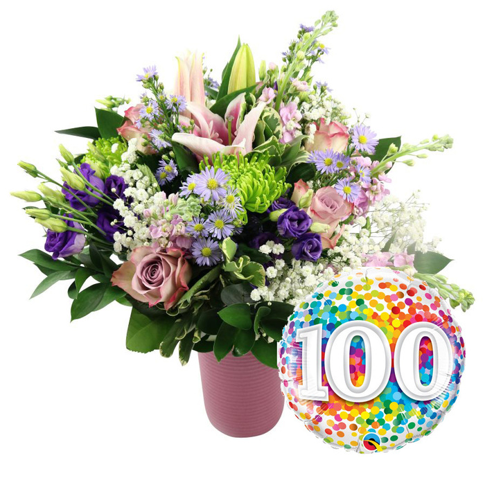 Set against a flawless white background, this image showcases a jubilant 100th-milestone bouquet designed with the refined eye of a Stanmore florist. A cylindrical vase in muted, matte pink with soft ribbing stands solidly at the base, its understated elegance allowing the lavish flowers above to shine. The bouquet rises in a loose, rounded shape, dominated by tall stems of light pink Oriental lilies that arc gracefully upward. Some lilies are wide open, displaying star-shaped petals with slightly darker pink veining and long stamens tipped with pollen, while others remain in smooth buds, promising a continuous unfolding of beauty through the celebration. Encircling the lilies are clusters of roses in complementary shades: blush pink for softness and deeper mauve or dusty rose for richness and depth. Their velvety petals are tightly layered, creating a sense of abundance and longevity. Between these roses, rich purple lisianthus petals curl and ruffle, adding a luxurious texture. Delicate lavender asters with sunny yellow centres sprinkle light throughout the design, echoing the informal charm of local Stanmore gardens. Bright lime green button chrysanthemums sit like tiny orbs, their textured surfaces catching the eye and bringing freshness. Wisps of white baby's breath drift through the bouquet, creating a gauzy, cloud-like effect that lightens the entire composition. Various dark green leaves, from broad and glossy to slender and arching, frame the flowers, giving the arrangement a full and balanced silhouette. To the lower right, overlapping the vase slightly, gleams a round foil balloon decorated all over with colourful confetti dots in red, orange, yellow, green, blue, and purple. At its centre, the large white numerals 100, with a subtle 3D shadow, stand out boldly, unmistakably marking a century of life or partnership. The whole scene radiates joy and gratitude, ideal for commemorating a 100th birthday, golden community events, or a centenary wedding anniversary in Stanmore.