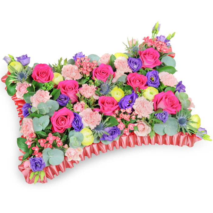 From a soft, oblique overhead perspective against a stark white background, this rectangular floral pillow reads as a carefully constructed field of blooms, dense and intentionally layered. The composition is dominated by hot-pink roses-some in tight bud, some opening into full velvety cups-whose saturated colour anchors the design. Surrounding them, pale pink spray carnations add ruffled volume and an almost textile-like softness, while purple lisianthus contribute deeper tonal pools and graceful bell shapes. Bright yellow ranunculus, their many delicate petals forming buttercup-like rosettes, inject cheerful accents that lift the palette away from strictly somber tones. Blue-silver Eryngium thistles punctuate the surface with spiky geometry and a cooler metallic note, a textural contrast to the smoother petals. Interspersed filler blooms in soft pink and creamy tones add breath and detail, and rounded eucalyptus leaves with their characteristic silvery sheen provide a calming, aromatic backdrop. The entire arrangement is edged by a deep red, pleated satin or silk ribbon-the ruffled border crisply defines the pillow silhouette and lends a refined finish. The lighting is even and natural-feeling, highlighting petal textures and the subtle sheen of the satin trim. As a sympathy cushion intended for memorial display-perhaps beside a service at a local Stanmore church or laid at a graveside in Stanmore Park-the piece speaks of remembrance with warmth and colour, offering solace through its joyful yet respectful combination of flowers and foliage familiar to families across the Stanmore Broadway area.