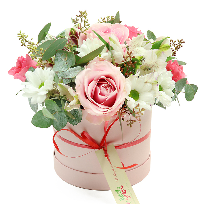 Centred against a pristine white background, a pastel pink hatbox overflows with a sumptuous bouquet, the kind of design you might have delivered to a Stanmore home overlooking the green spaces near Canons Park. The cylindrical box is finished in a matte blush tone, calm and refined, and is cinched with a narrow coral-red satin ribbon that runs neatly around the circumference. At the front, the ribbon is tied in a tidy bow, from which a cream-coloured rectangular tag hangs, bearing soft green script and a small website address that mark this as a curated gift piece rather than a simple shop posy. Above the rim, the flowers rise into a well-rounded dome, with no gaps or harsh lines, just a continuous sweep of colour and texture. A large bi-colour rose, fully opened, sits prominently near the front; its petals move from pale, almost ivory-pink edges into a deeper pink core, creating a natural focal point. Surrounding it are clusters of crisp white daisy chrysanthemums, each bloom centred with a bright green eye that adds freshness and contrast. White alstroemeria blooms, slender and slightly flared, are scattered throughout, their petals etched with fine speckles. Nestled between these are smaller, vibrant pink flowers-likely spray roses or lisianthus-that introduce rich, saturated colour notes. The bouquet is generously framed with greenery: rounded, silver-green eucalyptus leaves give a cool, contemporary edge, while darker green foliage and delicate stems with tiny green buds add both height and depth. The lighting is soft and even, revealing the subtle variations in petal colour and the velvety textures of the roses. Overall, the arrangement feels romantic but not overly dramatic, ideal for marking birthdays, engagements, or quiet celebrations with same-day delivery across Stanmore, from Stanmore station to the peaceful residential roads off The Broadway.