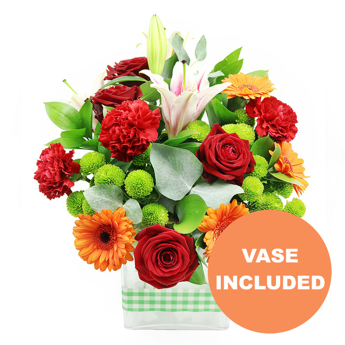 A colourful, densely packed bouquet bursts from a clear rectangular glass vase, its vivid tones and textures standing out sharply against a pure white background, like a centrepiece set for a family celebration in a Stanmore home. The vase is wrapped at the base with a light green and white gingham ribbon, its soft fabric adding a rustic, comforting note to the modern glass form. Dominating the central area are several deep crimson red roses, their petals tightly furled in rich, velvety spirals that suggest depth and enduring affection. Interlaced with these are bright orange gerbera daisies, each bloom showing a ring of flat, evenly spaced petals surrounding a dark brown central disc, creating an energetic focal contrast. Among them, red-orange carnations with heavily ruffled petals introduce a frilled texture that makes the bouquet appear lush and full. Rising above the main mass of colour, white oriental lilies add height and elegance; some are fully open, revealing long, trumpet-shaped petals subtly streaked with pink along their centres and prominent stamens dusted with pollen, while others remain closed in elongated buds, hinting at further beauty yet to unfold. Throughout the arrangement, compact clusters of lime green pompom chrysanthemums add a lively, almost playful element, their spherical heads and crisp hue giving the design a contemporary edge. Framing the blooms is an abundance of greenery: broad, deep green leaves provide structure and depth, while soft, silvery eucalyptus stems weave through, their muted tone and gentle arcs creating a natural, slightly airy outline reminiscent of walks through the green spaces around Stanmore Country Park. In the bottom right corner, a bold orange circular badge overlaps the vase and stems, with the clear white words VASE INCLUDED, signalling that this is a complete, ready-to-display bouquet, ideal for same-day delivery in and around Stanmore for birthdays, anniversaries, or heartfelt congratulations.
