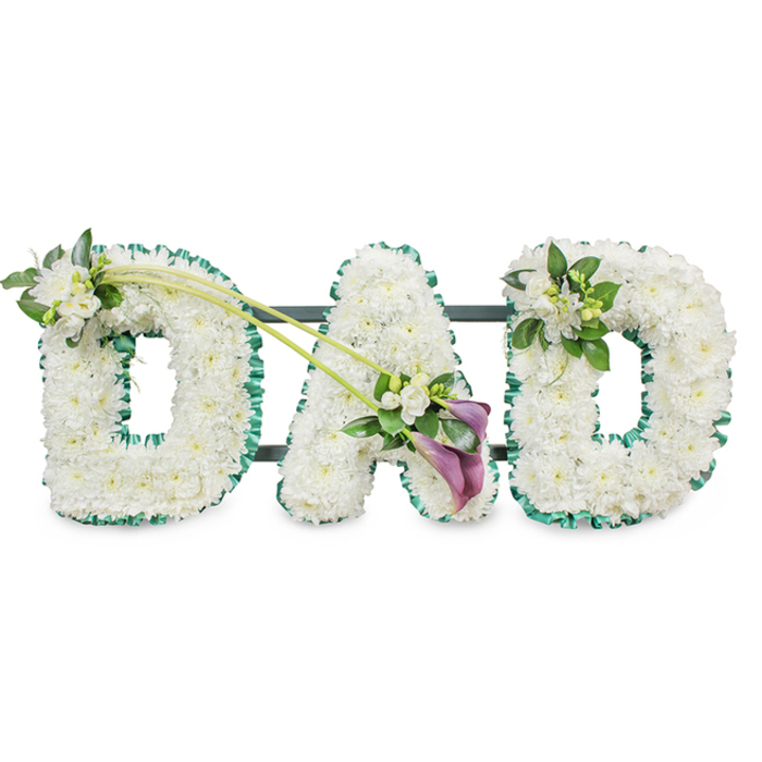 Captured against a bright white background, this image features a personalised funeral tribute spelling out DAD in large, three-dimensional floral letters, created with the kind of care you might expect from a dedicated Stanmore florist. Each letter is meticulously covered with small white chrysanthemums, packed closely together so their rounded, daisy-like heads form a smooth, continuous surface. The pristine petals overlap slightly, catching the light in gentle highlights while soft shadows settle in the spaces between, giving the arrangement a plush, cushion-like appearance that feels comforting and serene. Around the perimeters of the letters, an emerald green satin ribbon is carefully ruffled and pinned, hugging every curve and edge to define the shapes with crisp precision. This border adds a touch of richness and mirrors the verdant feel of Stanmore's tree-lined avenues and green open spaces. Subtle glimpses of dark structural bars behind the letters reveal the sturdy frame that allows the piece to stand upright during a service or graveside committal.
Thoughtful accent flowers bring individuality and emotional nuance to the design. On the first D, a dainty arrangement of white freesia blooms and polished green leaves sits at the upper left curve. Some flowers are fully open, while others remain in bud, suggesting ongoing love and memories that continue to unfold. From this focal point, two long, slender green stems sweep diagonally across the composition toward the central A, visually binding the letters together like a ribbon of shared family history. At their meeting point lies an elegant, slightly off-centre spray composed of two deep purple calla lilies surrounded by more white freesia and lush foliage. The calla lilies' velvety, trumpet-shaped blooms add a note of depth and gravity, their darker hue standing out against the white without overpowering it. On the final D, a smaller echoing cluster of freesia and foliage rests at the top right, completing the visual rhythm. Overall, the tribute conveys quiet strength, enduring affection, and gratitude for a father's presence, a fitting floral memorial for families in Stanmore seeking a meaningful way to say goodbye.