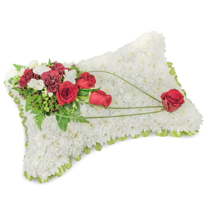 A refined and sorrowful floral tribute shaped like a rectangular pillow, photographed on a neutral white backdrop to emphasise its calm presence. The surface is completely built from hundreds of compact, snow-white chrysanthemum blooms, their layered petals forming a dense, cushion-like texture that suggests purity, rest, and consolation. A discreet border of pale lime-green ribbon or narrow foliage peeks from beneath the flowers, giving the arrangement a neat, finished rim. On the upper left sits an artful spray of deep reds: velvet roses at various stages of bloom and frilly, dark burgundy carnations, contrasted by small groups of delicate white blooms - likely freesias or small spray chrysanthemums - that add lightness and subtle perfume. Rich green foliage, including feathery fern fronds and sturdier laurel-like leaves, provides depth and supports the floral cluster. From this corner cluster, three graceful arcs of bright green bear-grass or similar linear foliage extend diagonally toward the lower right, creating movement and connection across the white plane; these lines culminate in a single, perfectly formed scarlet rose resting near the lower right edge, a poignant symbol of love and remembrance. As a classic bereavement piece often chosen for funerals or memorial services, the cushion speaks plainly yet tenderly - its combination of white chrysanthemums, red roses and carnations, and fresh green accents conveys respect, enduring affection, and a serene farewell suitable for families in Stanmore and surrounding neighbourhoods.