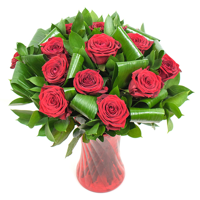 A richly detailed study of classic floral form: a compact, rounded cluster of more than a dozen deep crimson roses, each blossom fully open with densely layered, velvety petals that unfurl from a tight spiralled heart. The roses' colour is saturated and sumptuous, a true romantic red that reads almost jewel-like beneath gentle, uniform lighting. Surrounding the clusters is a volume of varied green foliage - smaller, glossy leaves create a steady, verdant bed while larger, broad leaves have been skillfully folded and rolled into elongated cylinders and dramatic curls, introducing an architectural motif that contrasts the soft organic curves of the roses. The craftsmanship is evident in the thoughtful placement of each leaf and bloom, producing visual breath and support without overwhelming the central roses. The bouquet is anchored in a tall, translucent ruby-red glass vase with a refined ribbed texture and a slight waist that lends the arrangement an elegant silhouette; the glass catches light in vertical streaks that mirror the stems and intensify the red harmony. Against a pure white backdrop, every nuance of texture - the velvet of petals, the gloss of foliage, the glass's faint swirl - is amplified, inviting the viewer to note the depth, fragrance, and tactile promise of fresh blooms. This composition is ideal for romantic milestones, formal centrepieces, or a polished gift for someone in Stanmore, whether placed on a mantel after a visit to Stanmore Common or chosen as a thoughtful anniversary bloom.
