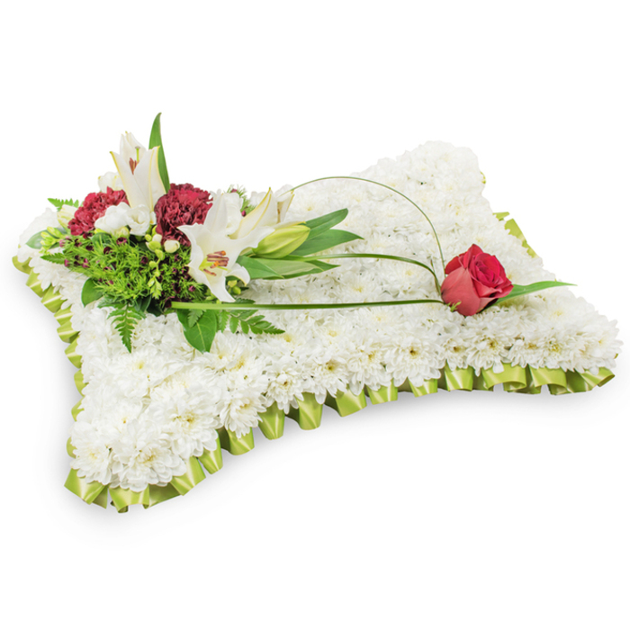 Presented against a simple white background, this classic funeral pillow is built from a dense, even field of small white chrysanthemums whose tight petals create a smooth, plush surface that speaks of purity and peaceful rest. A neat pleat of pale lime-green satin ribbon edges the rectangle, its delicate sheen and soft folds adding a refined finishing touch reminiscent of carefully laundered linens. At the top left a sculpted cluster forms the arrangement's focal point: two white Oriental lilies stand open with bright yellow stamens beside a closed lily bud, while deep burgundy spray carnations nestle among them to introduce rich, contemplative colour. Layered greenery - feathery ferns, broad-leaved salal and other glossy foliage - frames and supports the flowers, creating shadow, depth and a sense of living calm. From that cluster a slim arching strand of green leaf or decorative wire sweeps gracefully across the white cushion and terminates in a single deep red rosebud on the right, poised and just beginning to unfurl as a symbol of enduring affection and remembrance. The interplay of textures - the chrysanthemum's soft cushion, the lilies' smooth petals, the spray carnations' ruffled hearts and the ribbon's satiny edge - reads like a quiet, carefully composed poem of condolence. Crafted with local care by Flowers Stanmore and suited to display at a funeral, memorial or wake near Stanmore Tube or Bentley Priory, this piece conveys heartfelt sympathy and honours a loved one with traditional dignity.