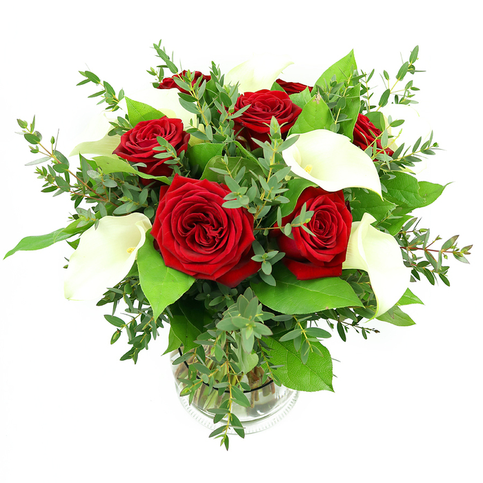 A sophisticated bouquet of red roses and white calla lilies is artfully arranged in a clear, cylindrical glass vase, photographed against a clean white backdrop that emphasizes its rich colours and textures. At the centre of the design, six or seven red roses are in full bloom, their densely layered, velvety petals curling inward in deep crimson spirals that exude warmth, devotion, and classic elegance. Threaded among these are graceful white calla lilies, around four to five stems, each one a sculptural trumpet of smooth ivory that curves gently upwards, revealing a slender yellow-orange spadix that adds a subtle focal point within the bloom. The interplay of bold red and tranquil white creates a sense of balance that feels both powerful and serene. Surrounding the central flowers is a generous assortment of greenery: fine stems of eucalyptus with soft, rounded, silvery-green leaves bring a delicate, airy movement, while broader, darker green foliage forms a solid base, offering structure and a lush, verdant frame for the brighter blossoms. The transparent vase holds the neatly cut stems in clear water, and a lightly ribbed band around its base provides a discreet decorative detail without competing with the flowers. Bright, neutral lighting illuminates the arrangement from the front, highlighting the velvety texture of the rose petals, the smooth sheen of the calla lilies, and the varied tones of the foliage, without harsh shadows. The overall impression is of a luxurious yet refined floral display, ideal as a centrepiece in a Stanmore home overlooking Bentley Priory Nature Reserve or as a romantic delivery from Flowers Stanmore to a townhouse near Stanmore station, perfectly suited for anniversaries, proposals, or any special occasion where heartfelt sentiment and polished style are desired.