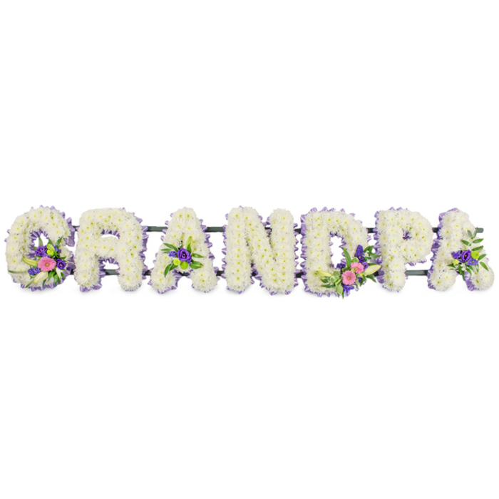 A beautifully composed funeral flower display spells out GRANDPA in bold, upright letters, designed as a personalised tribute for a much-loved grandfather. Each letter is made from a densely packed cluster of small white chrysanthemums, forming a plush, cloud-like surface that flows seamlessly from one petal to the next. There are no gaps in the coverage, giving the impression of letters carved entirely from white petals, all glowing softly under even, neutral lighting. Surrounding each letter is a fine trim of lilac and light purple blooms, possibly statice or tiny asters, which frame the white centres with a delicate, cool-toned outline. This subtle border adds clarity and definition to the lettering while keeping the overall mood gentle and respectful. Within the word, select letters carry extra floral accents that bring colour and character. The G features a small spray of bright pink flowers combined with a slender white lily and fresh green foliage, drawing attention to the beginning of the tribute. The first A and the final A both hold smaller clusters of rich purple flowers with deep green leaves, while the D showcases a more generous arrangement, echoing the G with vibrant pink blooms, another white lily, and verdant greenery. Here and there, small green button chrysanthemums or similar round blooms add a touch of brightness among the whites and purples. A slim, dark green frame runs along the back, giving the letters structure and helping them stand securely when placed at a service. Against a plain white background, the arrangement appears calm and composed, an elegantly traditional choice for a Stanmore family saying farewell to their grandfather, whether the service is held near Stanmore station, in a local church, or at a nearby crematorium. It expresses love, respect, and the quiet dignity of a life that has touched many others.
