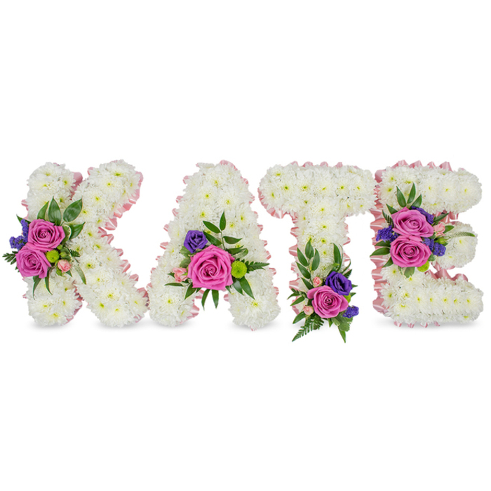 A serene and meticulously formed name tribute spelling KATE, each letter sculpted from a dense, snow-white mass of chrysanthemums that together create a soft, cloud-like texture and a strong three-dimensional presence against a clean white background. The edges of the letters are gently traced with a light pink satin ribbon, which reads like a blush outline and helps separate each character from the background while enhancing the piece's delicate mood. Bright accent clusters of flowers are placed with intentionality: bold magenta-fuchsia roses provide focal points, complemented by pale pink spray roses that add tenderness, and small deep purple or blue blooms-likely statice or lisianthus-introduce a refined, cool contrast. Floral placement is carefully considered for balance-on the K the diagonal stroke is brightened by a cluster; the A receives a spray near its lower right; the T is anchored with a grouping at its base; and the E bears two floral accents on its middle and bottom bars. Intervening foliage, including airy fern fronds and smooth eucalyptus leaves, offers a green counterpoint, subtle texture, and the faint, calming scent of fresh cut greenery. The arrangement is captured in soft, even light that emphasizes texture and colour without harsh shadows, creating a composition that reads as both dignified and consoling. Intended as a sympathy or memorial display, this bespoke floral lettering embodies the tenderness of remembrance, making it a fitting tribute for services in Stanmore, from small chapel gatherings to graveside ceremonies near Stanmore Common or local parish churches. The workmanship reflects experienced floristry, shaping emotion into form.