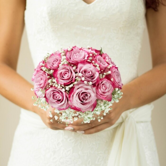 This close-up, poised in the heart of Stanmore, captures a moment of bridal anticipation-a bride's hands tenderly holding a round bouquet against her ornate gown. The arrangement is precision-crafted: medium-sized roses unfold with petals gently blushing from pale pink at their core to saturated fuchsia tips, forming a sphere of colour and lushness. Threads of delicate white baby's breath interlace the bouquet, their airy presence reminiscent of early morning mists settling over Stanmore Common. Foliage peeks out discreetly, suggesting careful hand-tying just beyond view. The bride's manicured nails hint at understated glamour, her skin warmly illuminated, while the elegant wedding dress-floral lace over the bodice, a soft V-neck, and a flowing sash-recalls ceremonies at Stanmore's historic churches. The soft, light background keeps every eye on the bouquet, amplifying its role as a symbol of joy and love. This image, created by Flowers Stanmore, celebrates the emotional resonance and attention to detail expected from a treasured local wedding bouquet, perfectly suited for memories made in Stanmore's charming community.