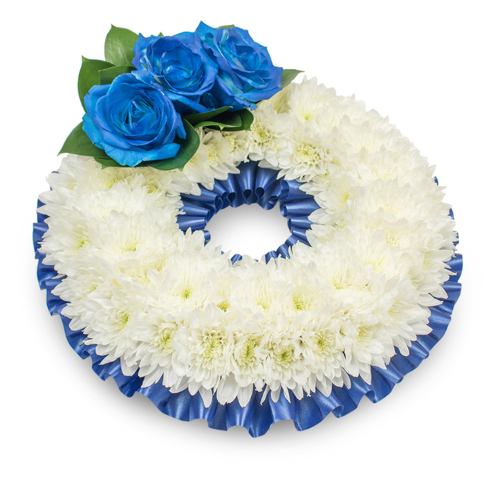 Viewed from a slightly elevated perspective against a pristine white setting, this memorial wreath is an eloquent composition of white and blue, shaped by careful floristry and thoughtful intent. The principal mass is a compact, slightly domed ring of immaculate white chrysanthemums; their numerous, delicate petals overlap in a soft, feathery texture that catches the light like powdered snow and conveys a sense of purity and calm. Positioned at the upper-left sector are three intensely blue roses, their petals richly hued and velvety, arranged to form a small, dignified focal cluster. Beneath those roses, dark green foliage provides a natural cushion and visual counterpoint, making the blues read deeper and the whites read purer. A pleated satin ribbon in complementary royal blue trims both the inner and outer circumferences, its glossy ruffles lending structure, sheen, and a finished, ceremonial edge to the piece. The gentle shadow beneath the wreath anchors it to the surface and suggests quiet gravity; the subtle scent of chrysanthemums mixes with the faint, deeper aroma of roses to create a restrained, comforting ambience. As a florist who often prepares tributes for families in Stanmore, I imagine this wreath placed with dignity at a chapel in the area or laid at a graveside near Canons Park - a composed offering that communicates respect, solace, and enduring remembrance through its textures, hues, and craftsmanship.