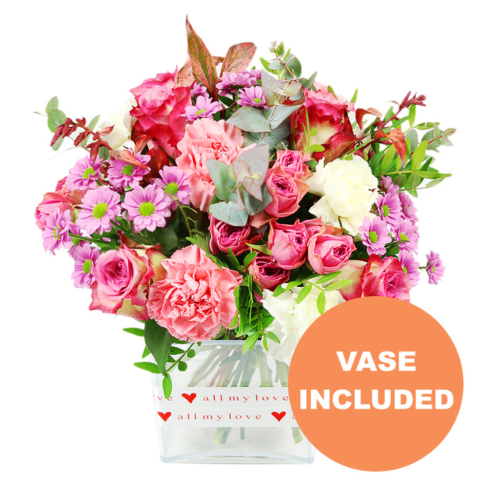 Centred against a pristine white studio backdrop, this photograph showcases a lush, carefully balanced bouquet brimming with pink blooms and soft greenery, as if freshly arranged in a Stanmore florist's workshop. Dominating the composition are multiple large roses, some fully open and others still unfurling, in rich fuchsia, magenta, and gentle blush pink tones. Their petals appear thick and velvety, curling elegantly towards the edges. Nestled between the roses, ample carnations in coral, salmon, and pastel pink provide a dense, frilled texture, filling out the shape of the bouquet and catching light in their ruffles. Dotted among these are daisy-like chrysanthemums in shades of lavender and purple, their bright green central discs creating sharp, cheerful contrasts. Creamy white flowers are subtly interspersed, adding clarity and lifting the overall palette. The arrangement is framed and threaded through with abundant foliage: rounded eucalyptus leaves in silvery green, mixed with darker, glossy leaves and a few touches of reddish-brown greenery that lend a hint of warmth, almost like the turning leaves of autumn around Stanmore Common. All the stems are gathered neatly into a clear square glass vase, whose transparency reveals the lower portion of the stems and water line, reinforcing the sense of freshness. Around the base of the vase, a crisp white decorative band is wrapped, printed with repeating red hearts and the handwritten phrase all my love, making it unmistakably a gift of affection. In the lower right corner of the image, a bold orange circular badge with white capital letters reads VASE INCLUDED, drawing the eye and clarifying that the arrangement arrives ready to place directly on a table. This bouquet feels ideal for delivering heartfelt sentiments to homes along The Broadway, near Stanmore Golf Club, or tucked in the quiet residential streets of HA7.