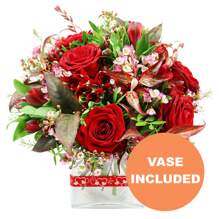 A richly coloured, romantic bouquet is presented here against a clean white background, as though ready to be carried through the streets of Stanmore to brighten a special evening. The focal point is a collection of several red roses, each bloom displaying layers of velvety petals in a deep, saturated hue. Some roses appear freshly opened, with tight, defined centers, while others have unfurled into broad, generous blooms that seem to spill over with colour and emotion. Interlaced with these are clusters of deep red flowers, resembling alstroemeria or similar multi-petalled varieties, which create pockets of texture and reinforce the passionate palette. Scattered delicately among the stronger reds are airy sprays of tiny pink and white blooms, echoing the look of waxflower; their minute, star-like petals and rounded buds offer a gentle, almost cloudlike lightness, preventing the arrangement from feeling too heavy. A generous collar of foliage surrounds the flowers, featuring glossy green leaves, lighter green sprigs, and textured stems that contribute depth and freshness. Standing out among the greens are dramatic burgundy and reddish-brown leaves, some subtly veined or marbled, which add a sophisticated, almost dramatic undertone that feels fitting for milestone celebrations, from anniversaries to romantic birthdays. The bouquet has been shaped into a rounded, domed form, creating an impression of abundance and careful balance. It is set into a clear, contemporary glass vase with a rectangular base, the visible stems and clear water suggesting a bouquet freshly prepared and ready for delivery across Stanmore, from quiet residential lanes to busy local shops. Around the upper section of the vase, a red ribbon printed with repeating white hearts is tied smoothly, underscoring the romantic theme. In the lower right corner of the image, a prominent round orange label reads VASE INCLUDED in bold white capitals, clearly signalling that this striking arrangement arrives as a complete, display-ready gift.
