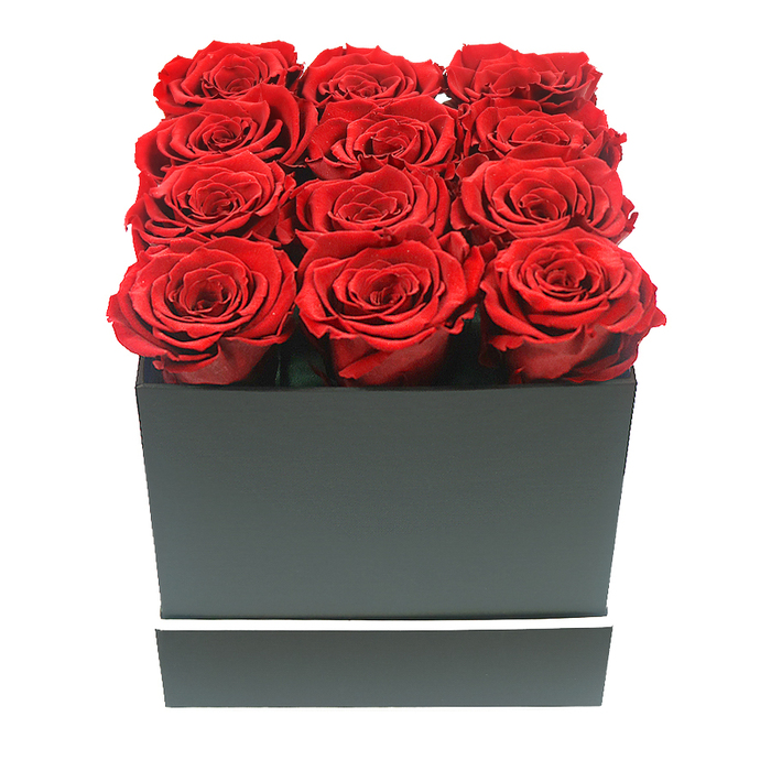 In this refined, studio-style image, a luxurious arrangement of twelve deep red preserved roses sits proudly in a sleek square box, the kind of thoughtful gift often ordered for special occasions in Stanmore. The roses are the undisputed stars: each bloom is fully open, with velvety petals tightly layered into lush spirals that create a sense of volume and softness you can almost feel. Their colour is a saturated ruby red with gentle variations that catch the soft lighting, shifting from darker wine tones within the petal folds to brighter crimson along the outer edges. Arranged in a precise 3x4 grid, the rose heads are nestled side by side so that they form a near-perfect rectangle of red, rising just above the rim of the container and giving the impression of abundance and care. The box itself is a matte black cube with smooth faces and sharp, defined corners, stylistically similar to the modern interiors seen in new apartments near Stanmore Station. A slim, clean white band encircles the lower part of the box, offering a simple, elegant contrast that emphasizes the graphic interplay of dark and light. The background is entirely white, allowing the arrangement to stand alone without distraction, while even, diffused lighting enhances the petals' velvety texture and casts delicate shadows along the box edges. From the perspective of an experienced Stanmore florist, this preserved rose design radiates modern romance and understated luxury-perfect as a long-lasting anniversary gift, a Valentine's surprise delivered to a home near Canons Park, or a chic decorative accent for a minimalist living room. The image conveys quiet intensity and enduring affection rather than fleeting bloom.