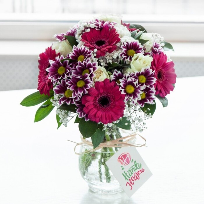 A beautifully composed mixed bouquet sits centre stage in a clear, subtly textured glass vase, set against a bright, softly diffused white background that could easily be a sunny Stanmore kitchen windowsill. The flowers are arranged with the practiced eye of an experienced local florist: striking gerbera daisies in deep fuchsia and magenta form the main focal points, their smooth, radiating petals framing dark, velvety centres that anchor the entire design. Surrounding them are generous sprays of bicolour chrysanthemums, each bloom showcasing fringed petals that start with intense purple at the tips and gently transition to crisp white, leading the eye towards warm yellow centres that bring energy and light. Nestled gently among these bold tones are creamy white roses, some still cupped in bud and others just beginning to unfurl, adding romance and softness as if they were chosen for an anniversary surprise sent across Stanmore and nearby Canons Park. Fine, cloud-like clusters of white gypsophila, commonly known as baby's breath, are threaded throughout the arrangement, giving it an airy, almost ethereal feel and breaking up the stronger colours with delicate texture. Rich, dark green foliage supports and frames the flowers, creating depth and a garden-fresh impression, as if the stems had been gathered at first light. The short, jar-like glass vase reveals neatly trimmed stems in clear water and is tied around the neck with a length of rustic twine, casually looped into a simple bow. From this bow hangs a small rectangular gift tag in soft white, adorned with a subtle pink floral graphic and fine lettering that suggests a handwritten note, even if the exact words cannot be seen. Lit by natural daylight, every detail-from the sheen on each petal to the gentle shadows on the vase-contributes to a feeling of freshness, care, and the quiet thrill of receiving a thoughtfully arranged bouquet from a trusted Stanmore florist.