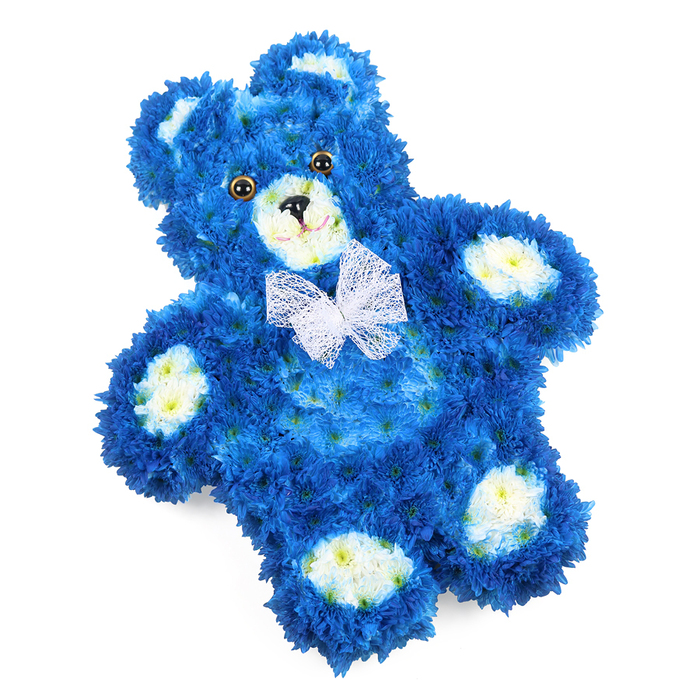 A three-dimensional teddy bear made entirely from blossoms rests on a bright white background, radiating both innocence and quiet reflection. The main body of the bear is composed of densely packed royal blue flowers, each miniature bloom stacked and layered so closely that the surface appears smooth, plush, and touchably soft, like the fur of a well-loved toy. The florist has carefully shaped every part-the rounded head, the gently curving arms, the slightly splayed legs, and the neat little paws-to capture the familiar teddy bear outline with lifelike precision. Accents of creamy white and pale yellow petals define the features: the inner circles of the ears, the rounded muzzle that pushes forward slightly, and the four oval paw pads that sit like soft footprints at the end of each limb. Set into the face are two dark, glossy eyes that lend a sense of character and presence, along with a small black triangle nose and a fine curved line suggesting a modest, comforting smile. Around the neck, a delicate white mesh bow is tied, its airy structure softening the strong blue colouring and introducing a ceremonial grace. The bear is photographed lying flat but positioned on a gentle angle, which enhances the depth and reveals the intricate petal work. In Stanmore, where families gather for farewells in local chapels and crematoria, or in quiet homes near Stanmore Country Park, this blue floral teddy is often chosen as a tribute for a baby boy or a loved one remembered for their childlike spirit. The deep blue tones offer a feeling of peace and reflection, while the teddy shape evokes protection, closeness, and unspoken love. Delivered with care across Stanmore, it becomes a tender, handcrafted symbol of remembrance.
