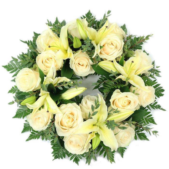 From an overhead perspective, this full, circular funeral wreath reads as a carefully balanced study in soft tones: cream-coloured roses and pale yellow lilies sit snugly within a rich, verdant bed. The roses - numbering around a dozen to twenty large blooms - show classic, tightly spiralled petals that glow with a tender, ivory warmth under soft, even lighting. Interspersed among them the lilies introduce vertical contrast; their trumpet-shaped petals unfold in pale lemon hues, some open to reveal slim, light-tinted stamens while others remain in elegant bud form, adding sculptural pauses across the surface. The greenery is deliberate and textural: fine, feathery fern fronds form a gentle halo, their filigree edges softening the wreath's outline, while broader sprigs of darker, rounded foliage, likely eucalyptus or similar, offer deeper green tones and a muted, leathery sheen. Placed on a clean white background, the wreath's circular geometry is clear and symbolically resonant, suggesting continuity and remembrance. The fragrance is understated - a powdery rose note supported by the fresh, green lift of lily and leaf - intended to soothe rather than startle. This arrangement is suited to funeral services, memorial tables, or a graveside setting and is composed with the restraint and dignity appropriate to such occasions. As a local Stanmore creation, it reflects a calm, respectful aesthetic that feels both traditional and gently contemporary for families seeking a composed, lasting tribute.