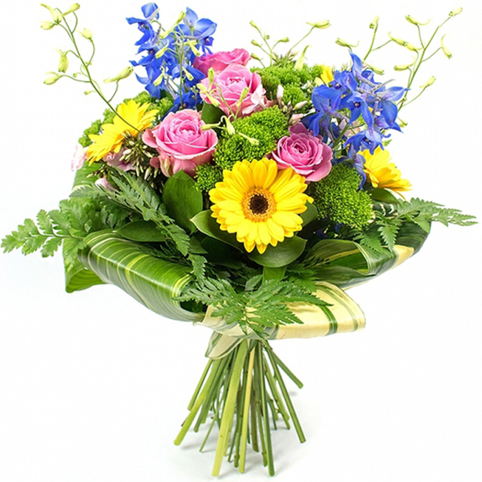This image shows a beautifully composed mixed bouquet standing tall against a pure white background, its colours and textures given full attention, as if you were viewing it in the calm light of a Stanmore living room. Several open pink roses sit at the heart of the design, their velvety petals forming loose spirals that feel both luxurious and inviting, suggesting love, gratitude, or gentle congratulations. Around this romantic core, bright yellow gerbera daisies with deep brown centres provide an instant burst of joy, their neat rows of petals radiating outwards like little suns, echoing the cheerful character of Stanmore's local cafés and friendly neighbourhood feel. Enhancing the overall shape, long stems of striking blue delphiniums rise gracefully above the rounded mass of blooms, each stem dotted with delicate florets that add movement and a cool note of refinement, a little like glimpsing a strip of clear sky between rooftops. The bouquet is wrapped in an abundant array of greenery: soft, feathery fern fronds arch delicately from the sides, giving a sense of lightness, while broad, polished tropical leaves are carefully curved and overlapped around the lower section of the flower heads. Some leaves are marked with subtle lighter stripes, which catch highlights and give a layered, botanical look. Between these, clusters of bright green, textured filler greens fill any spaces, adding volume and a slightly mossy, tactile feel. Fine, light-green stems with tiny unopened buds stretch subtly above the main flowers, reinforcing the idea of fresh, newly cut stems. At the base, the natural stems are gathered cleanly and bound with a soft cream or pale green wrap tied in a modest bow, leaving the ends exposed to emphasise the hand-tied craftsmanship. The overall impression is of a joyful yet elegant bouquet - perfectly suited to same-day delivery across Stanmore, whether to homes near Canons Park or offices around Stanmore Broadway.