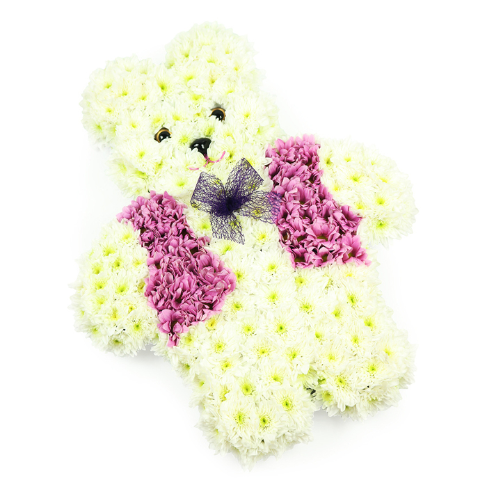 The photograph shows a floral teddy bear tribute designed with remarkable care, set against a clean white backdrop that lets every detail stand out. The bear's form is fully sculpted from white and creamy chrysanthemums, each flower nestled snugly into place to define the rounded head, small ears, curved arms, and solid little feet. The dense, cushion-like surface recalls the softness of a well-worn teddy, the sort of toy that might have once been cherished in a child's room overlooking the green spaces of Stanmore. Two small, glossy dark eyes are placed symmetrically on the face, giving the bear a tender gaze, while a neat black nose and a gentle curve of the mouth suggest a quiet, reassuring smile. Across the chest, a floral vest adds depth and contrast, created from blossoms in magenta, rose-pink, and lilac tones. These richer hues are carefully arranged to form two panels that lie like lapels over the lighter fur, lending the bear a dressed, almost ceremonial look. Just at the neckline, a sheer purple net bow is tied in soft folds, its translucent texture catching the light and introducing a whimsical, almost storybook charm. The lighting is even and soft, with minimal shadows, so the viewer can appreciate the three-dimensional modelling and the subtle variations of colour in each bloom. This floral teddy is the type of piece often chosen by families in Stanmore who wish to express deep affection and remembrance, whether for a child's service, a poignant anniversary, or a specially dedicated section of a funeral tribute. Prepared by Flowers Stanmore, it carries a sense of tender guardianship, as if standing watch beside a photograph or resting gently on a casket with love expressed in every petal.