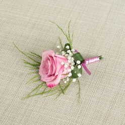 An elegant blush-pink rose boutonniere lies on a pale beige linen surface, the fabric's gentle weave visible beneath the floral work and calling to mind a carefully prepared dressing table in a Stanmore home before a special event. The single rose, caught at that perfect moment between bud and full bloom, shows a soft gradient of colour: a pale, creamy pink at its centre that gradually becomes a more saturated, romantic pink at the petal edges. Each petal is velvety and slightly curved, opening outwards in a calm, spiralling pattern that feels both classic and contemporary. Nestled at the base of the rose are clusters of white baby's breath, their tiny, star-like flowers creating a misty halo that lightens the composition and brings a traditional wedding charm often seen at ceremonies around Canons Park or Stanmore Country Park. Fine, slender strands of bright green foliage arc out from behind the rose, suggesting movement and giving the boutonniere a fresh, garden-picked feel, while a single, broader green leaf is folded and wrapped around the stems like a miniature cuff. The stems themselves are bound in a smooth, satin ribbon in shades of lavender and deeper magenta, tied in a neat knot with minimal trailing ends, adding a discreet touch of colour that gently echoes modern wedding palettes. Soft, diffused lighting from the side highlights the contours of the petals and the ribbon's sheen, creating subtle shadows that give depth without harshness. This carefully composed image evokes the quiet anticipation before a wedding or prom, when a finishing touch like this boutonniere is pinned to a lapel before stepping out into the streets of Stanmore for photographs and celebration.