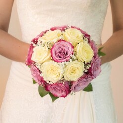 This evocative scene from a Stanmore bride's special morning encapsulates the detailed work and emotion behind every wedding bouquet carefully crafted in local workshops. The close-up reveals an exquisite round bouquet, each creamy yellow rose meticulously nestled beside fuchsia pink and muted lavender blooms, their satiny petals overlapping in a vibrant, textured tapestry. Fragile baby's breath threads between the blooms, adding white accents that resemble the early morning mist over Stanmore Common. Lush green leaves peek from the design's edges, offering a quiet contrast to the romantic hues. The bouquet is gently held by the bride, the purity of her white gown-crowned with lace embroidery-forming a perfect frame for the melodic clash of colours. Natural daylight brings fullness and clarity, making every flower seem almost touchable, while the softly blurred background ensures the arrangement remains centre stage. Thoughtfully styled by hands familiar with the tastes and traditions of Stanmore couples, this bouquet evokes love's quiet promise and the enduring artistry of Flowers Stanmore-a floral touch destined to become a cherished memory, perfect for wedding features or bridal inspiration.